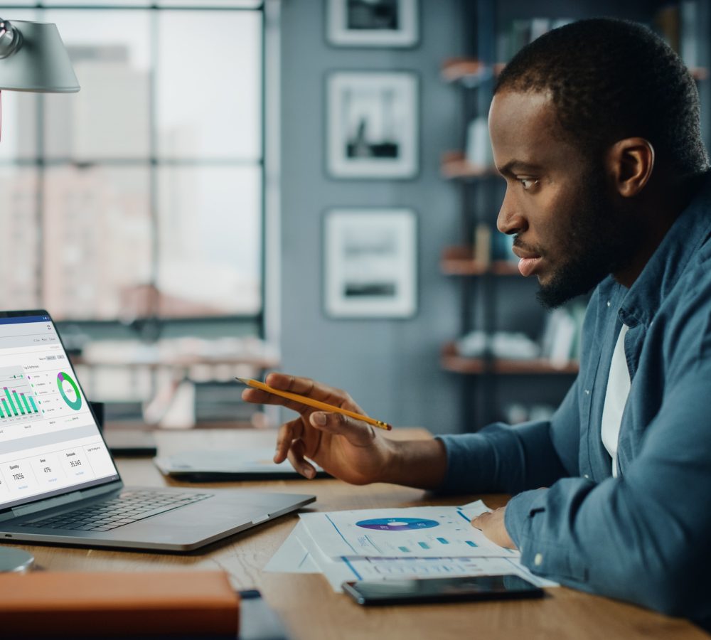 Handsome Black African American Specialist Working on Laptop Computer in Creative Home Living Room. Freelance Male is Doing Market Analysis and Creates Report with Charts for Clients and Employer.